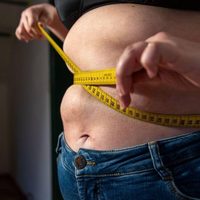 Eine Frau mit Übergewicht hält ein Maßband um ihren Bauch.