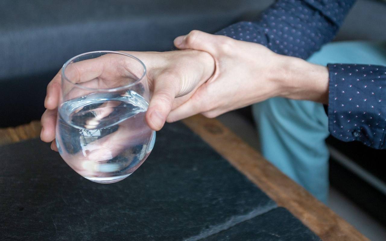 Mann hält seine zitternde Hand, die ein Wasserglas hält, fest