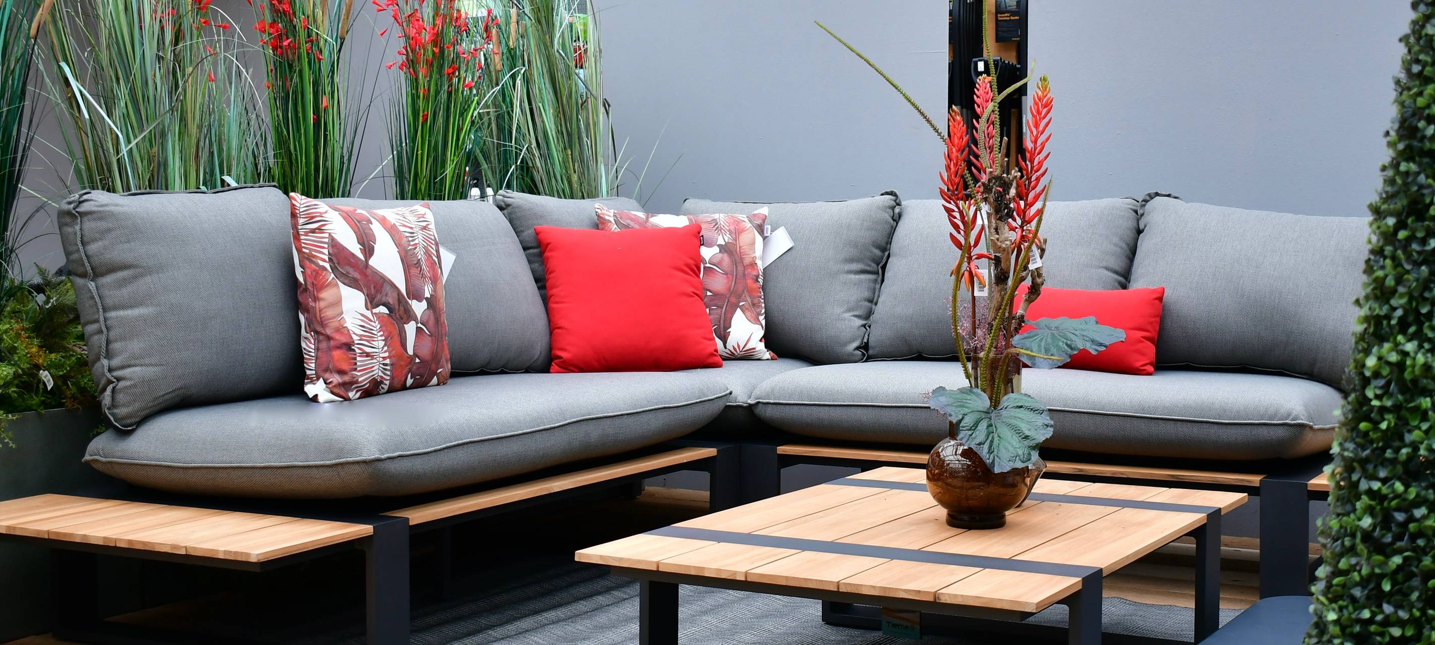 Auf dem Bild sieht man ein Sofa mit großen grauen und kleineren roten Kissen. Das Sofa hat eine "L"-Form. davor steht ein niedriger Tisch. Auf dem Tisch steht eine Pflanze in einer Vase.
